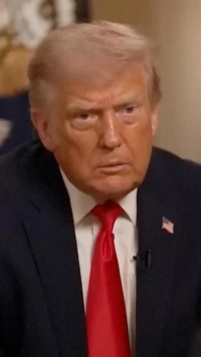 Donald Trump looks off camera as he sits down and wears a blue suit, red tie and American flag pin.