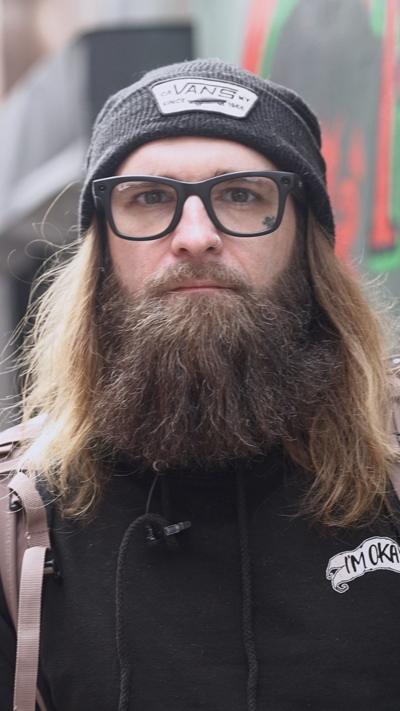 Tom is looking towards the camera. He is wearing a black hoodie, black beanie and black glasses. He is also wearing a light pinkish backpack. 