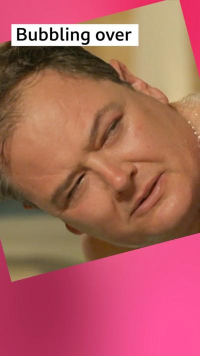 A man, Alan Carr squints. He is covered in foam, lying on a massage table.