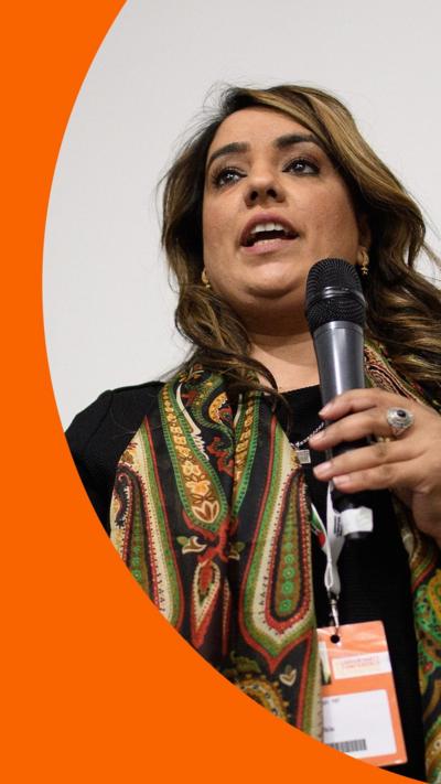Naz Shah is making a speech. She is wearing a black top and multi-coloured scarf, and holding a microphone.