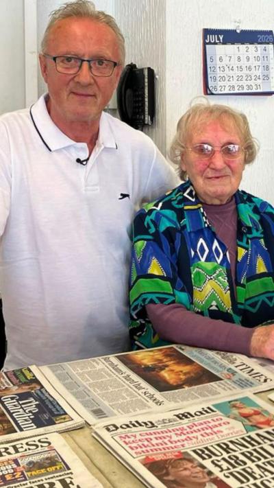 A man in a white polo shirt next to a woman in a green, blue and yellow shirt with purple top underneath. They are stood behind a newsagent's counter with newspapers on top