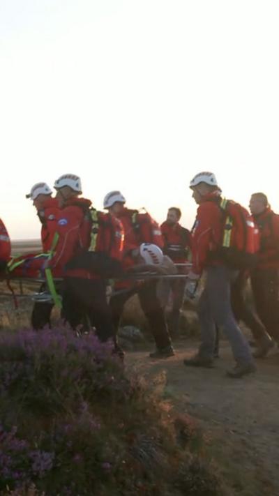 A group of people wearing red jackets and white hard hats carry a person on a stretcher over open moorland as part of a training exercise. The sky is bright with sunlight.
