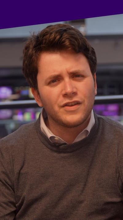 A man looks into the camera, he is wearing a grey jumper, with a blue collared shirt. 
