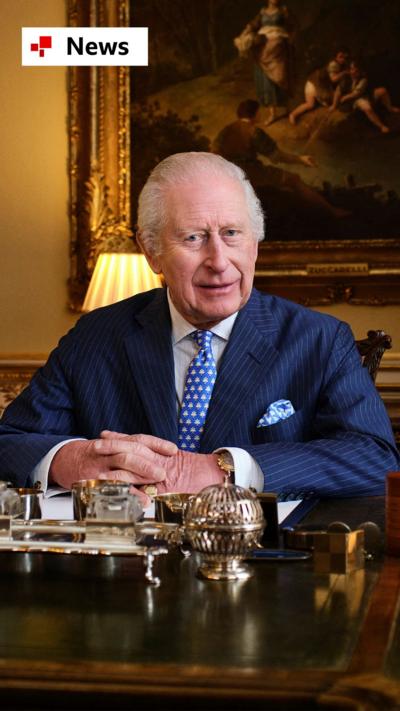 King Charles, wearing a suit, sitting behind a desk in Buckingham Palace with a painting behind