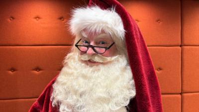 A man dressed as Santa Claus smiling to the camera with a long white beard, black glasses, rosy nose, a red and white Christmas hat and coat 