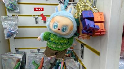 A fake Labubu hangs in a shop. It is blue and wearing a green jumper.
