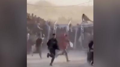 A crowd of people, one climbing over a fence, run to collect aid