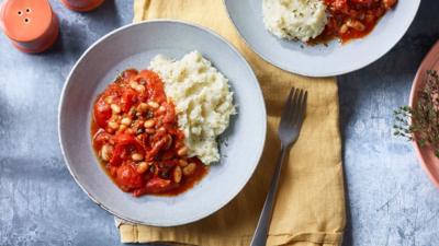 A light coloured plate with bean stew on one half and mashed cauliflower on the other sits on a yellow napkin with a fork by the side.