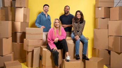 Four people - two women and two men - in casual clothes sit on cardboard boxes