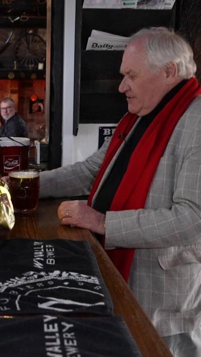 A man with grey hair at a pub bar. He is wearing a grey suit with a red scarf
