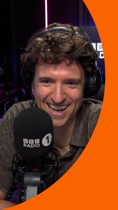 Greg James has wavy brown hair and is wearing a dark olive green shirt with a V-neck and collar.