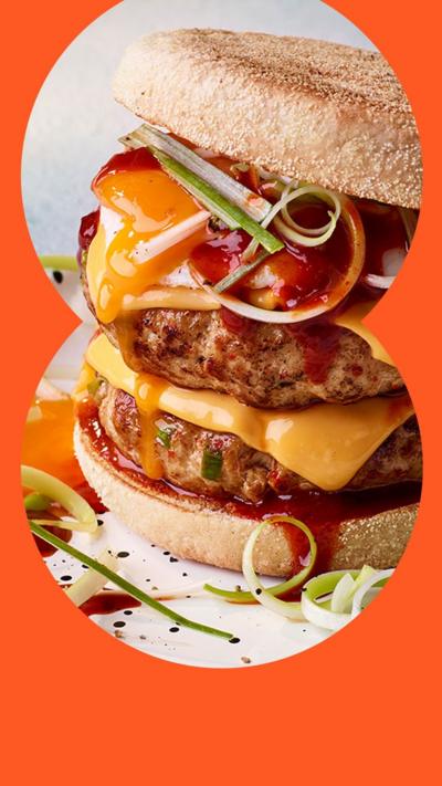 A close up shot of a breakfast muffin with two patties, a slice of cheese, veg and sauce