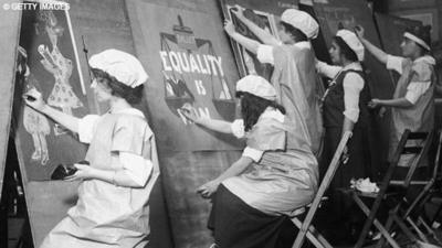 Young girls wearing aprons and caps make equality posters