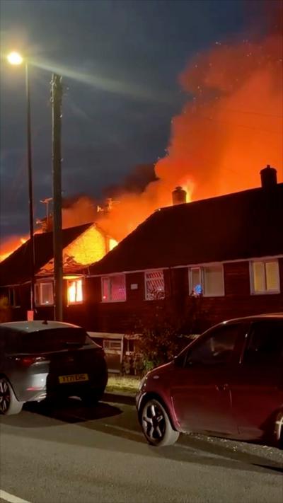 Smoke can be seen coming from behind a row of semi-detached homes in a residential street. It is dark. No flames are visible but the properties are illuminated by a fire burning behind them