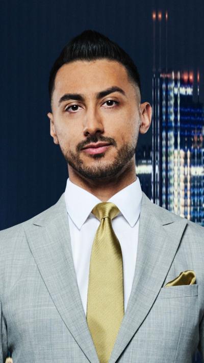 A young man with a beard wearing grey suit and gold tie