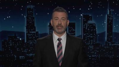 Jimmy Kimmel, wearing a dark suit and red-striped tie, speaks to the camera on his show.