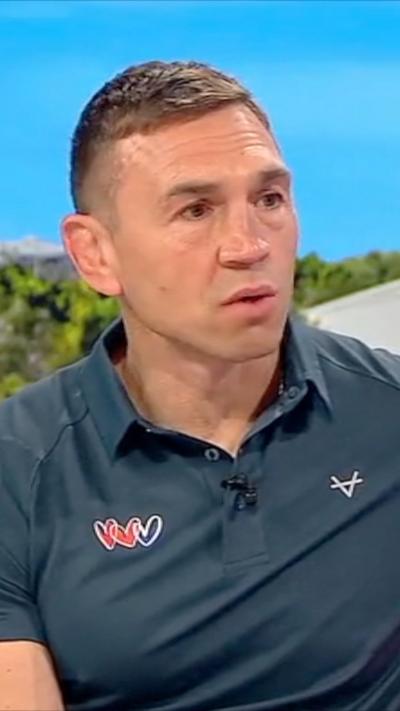 A man with short brown hair wearing a blue polo shirt sits on a red sofa in a TV studio