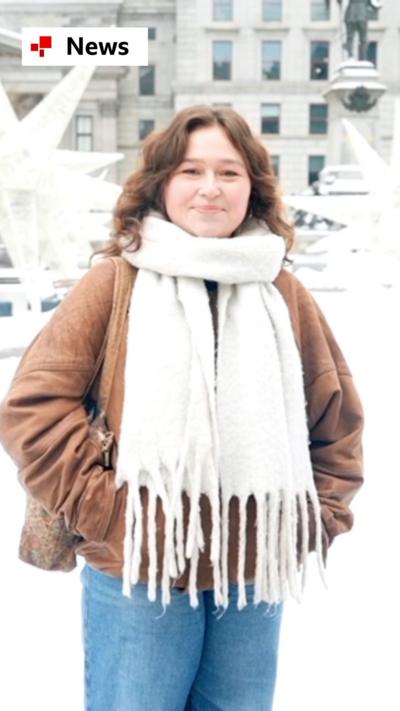 A young woman wearing a brown coat and white scarf speaking to the camera in a snowy square