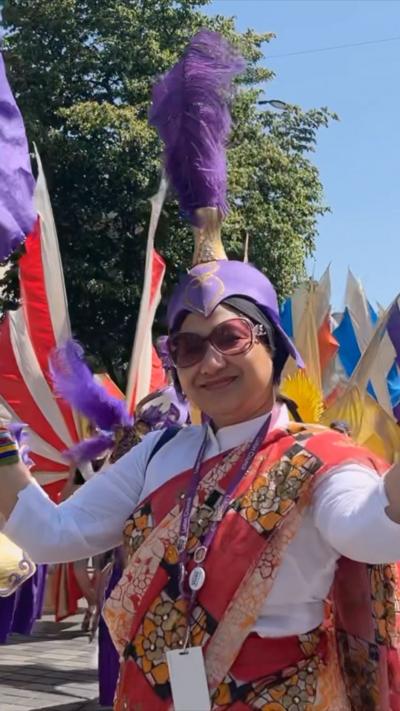 Person in a white top with an orange sharee-style fabric over the top. They are wearing a purple head piece with feathers