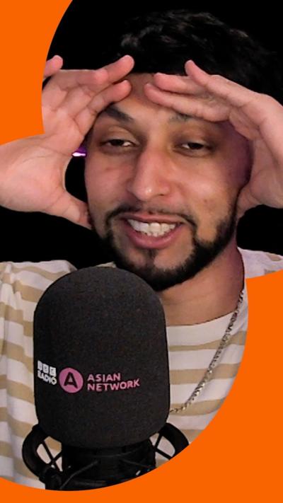 A man with short, dark hair and a short, dark beard holds his hands on his head as he speaks into a microphone.