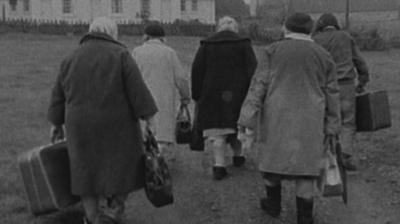 Five older ladies wearing long coats and carrying suitcases, walking 