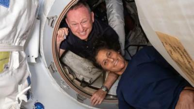 Butch Wilmore and Suni Williams onboard the ISS smiling as they pose in the orbiting lab. Butch is looking out from within a white circular hatch, while Suni is lying just outside it.