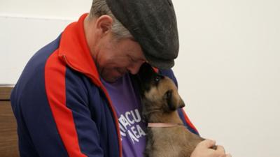 A man with a brown cap cuddling a brown dog. The man is wearing a blue and red tracksuit top and a purple t-shirt
