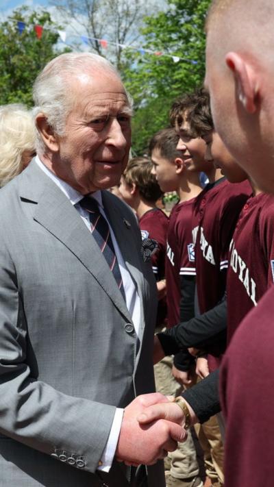 King Charles, wearing a grey suit, shakes the hand of a young man lined up in a row of other young men wearing burgundy t-shirts. Queen Camilla is behind him.