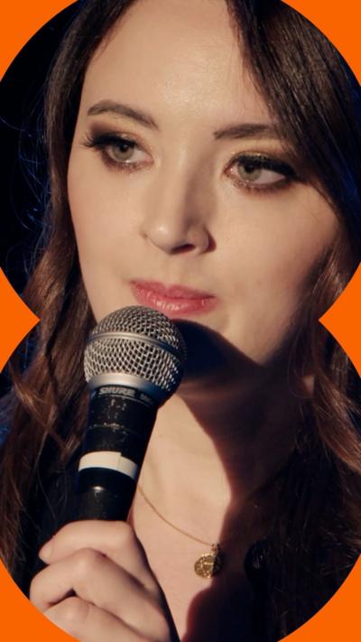 Fern Brady is stood on a stage, speaking to an audience with a microphone.