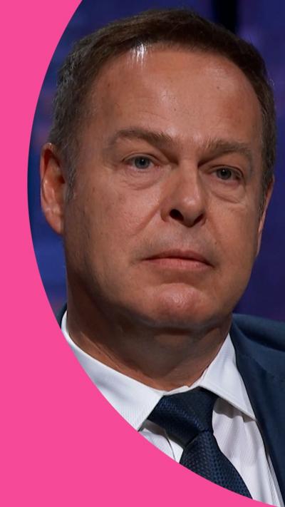 Peter Jones looks stony faced in the Den. He wears a dark blue suit jacket and tie, with a white shirt