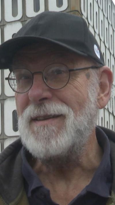 A man wearing round black frame glasses, a black cap and black polo. He has a grey beard.