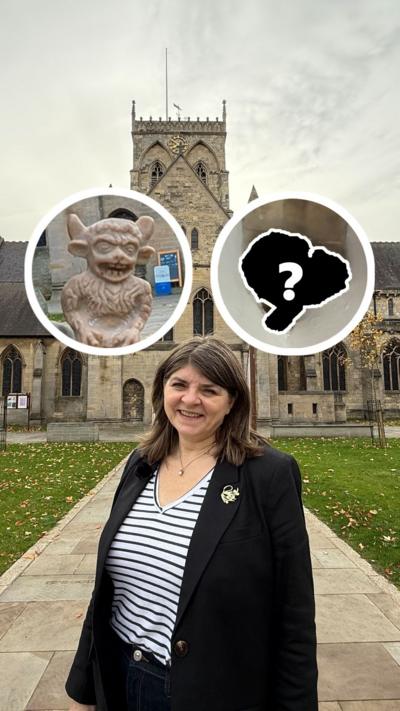 Emma Lingard has a long brown bob haircut and is wearing a black jacket and a black and white striped v- neck top. She stands in front of Grimsby Minster with graphics of the Lincoln imp and the Grimsby Imp next to her.