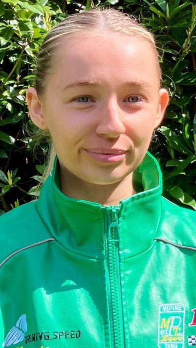 A close-up of Amie Donaghey. She is a young woman with slicked back, long blonde hair. She is wearing a green zip jacket and she is stood in front of a large bush.
