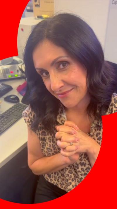 Presenter Maryam Moshiri smiles at her desk with her hands clasped together. She has long black hair and wears a leopard skin top