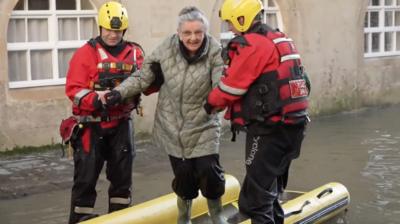 An elderly lady being helped out of a dinghy by two firemen in rescue gear