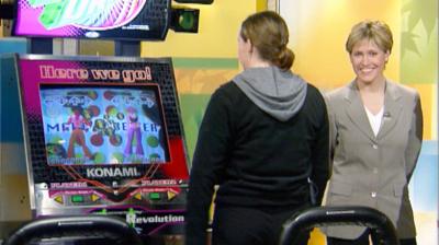 Woman plays dance arcade game while tv presenter looks on at the camera