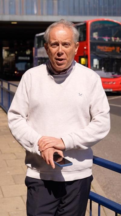 Reporter stands at bus station in Great Yarmouth, he has grey hair and is wearing a pale pink jumper and holding a mobile phone