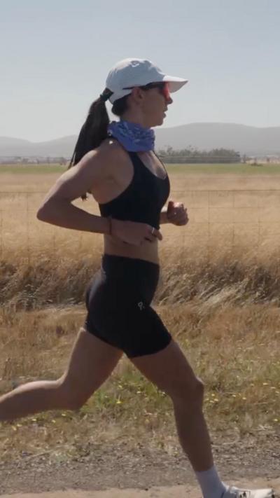 Woman wearing a white hat, neckscarf, sunglasses and a black running top and shorts running past a field