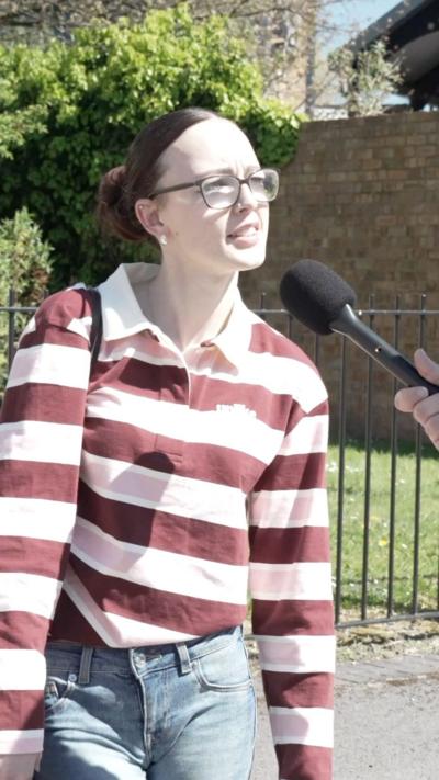 Woman speaking into a microphone wearing glasses, a red and white striped t-shirt and brown hair.
