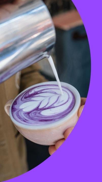 A hand holds coffee cup. Above it, milk is pouring from a jug to create foam art in a purple drink.
