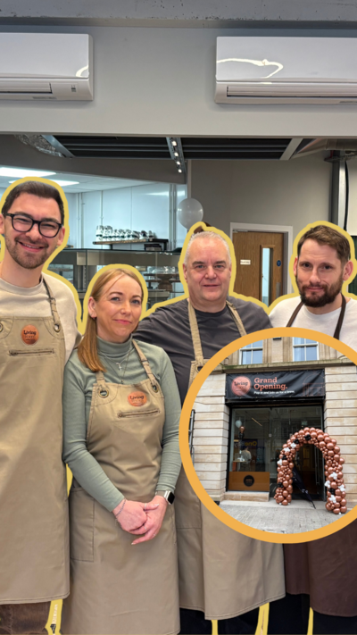 Four people - three men and a woman - stand in a cafe. They are wearing brown and beige-coloured aprons and are smiling into the camera.