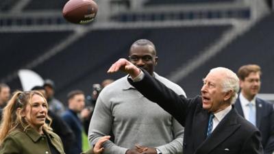 King Charles throws an American Football wearing a long coat and suit