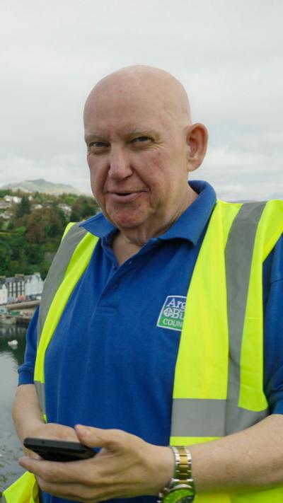 Colin Morrison wears a blue polo shirt and a hi-vis yellow vest. He is holding his phone in his hand and is smiling at the camera.