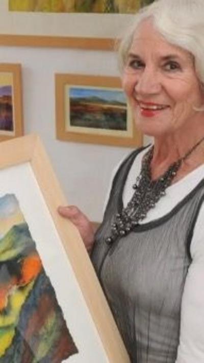 A woman with grey hair wearing a white top under a grey dress holding a painting