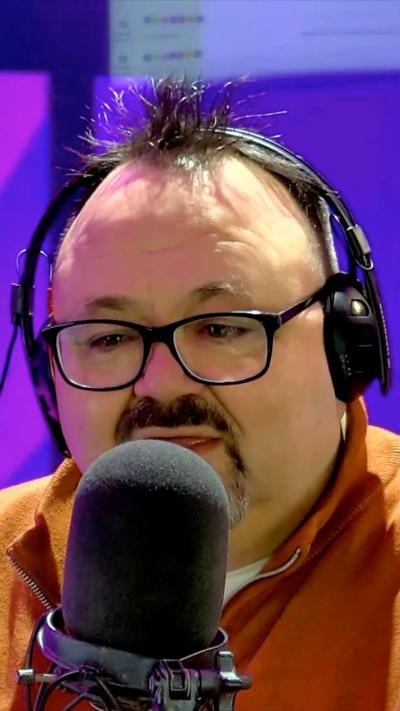 A man wearing dark-framed glasses and an orange top sits in front of a microphone with headphones on. Behind him there is a screen with a pink and purple pattern on it.
