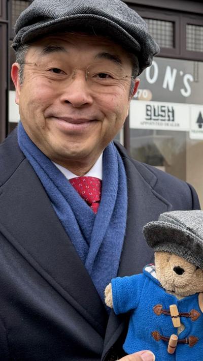 A Japanese man in a grey flat cap with blue coat, blue scarf, red tie and white shirt holding a paddington bear toy