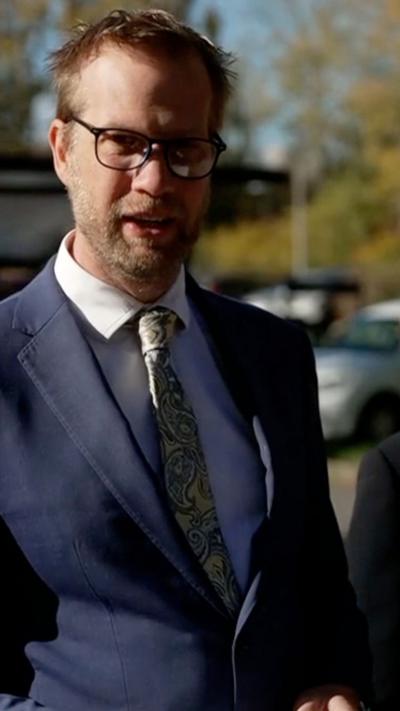 A man stood in a suit, wearing glasses, looking into camera
