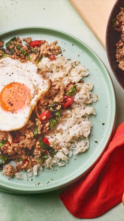 Top down view of stir-fried pork, rice and a fried egg on a green plate with cutlery next to it.