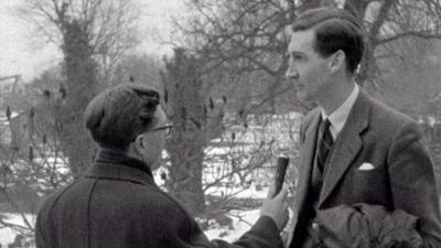 Black and white image of 91ȱ reporter on left holding a microphone in front of the local doctor.  In the back ground snow can be seen between the trees.