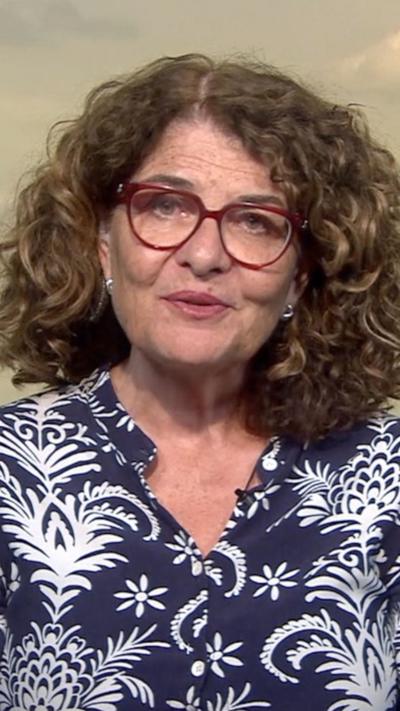 A woman with shoulder-length brown, curly hair and dark red glasses.